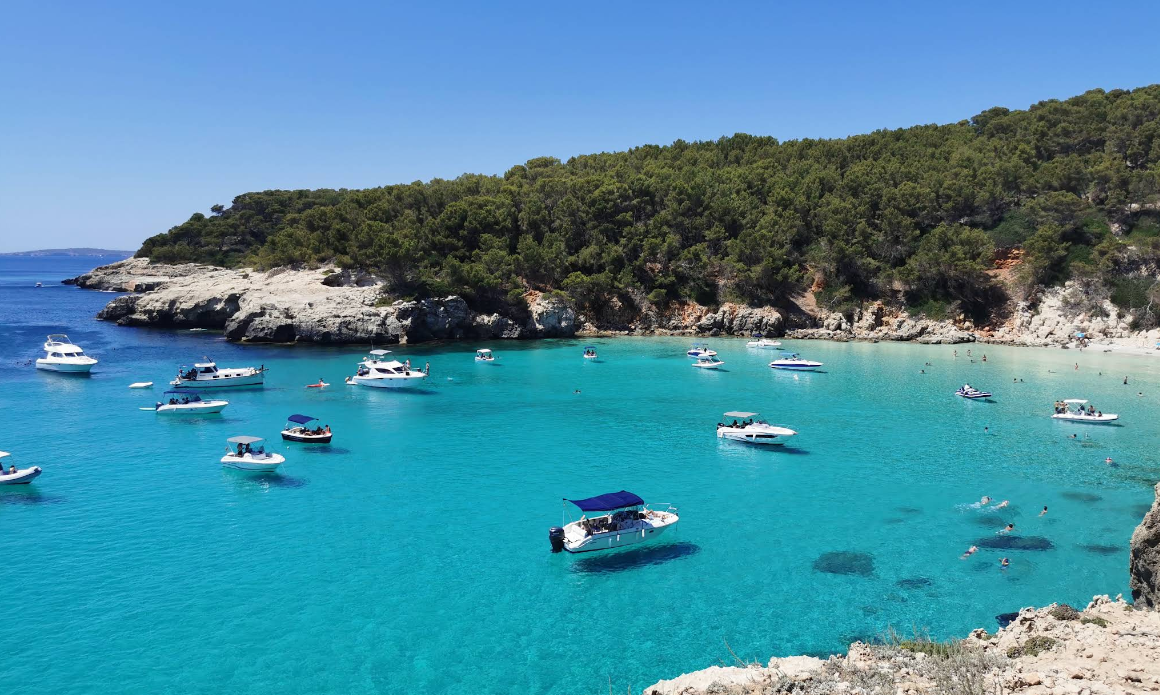 Cala Escorxada, a remote turquoise cove ringed by pine forest with boats anchored offshore