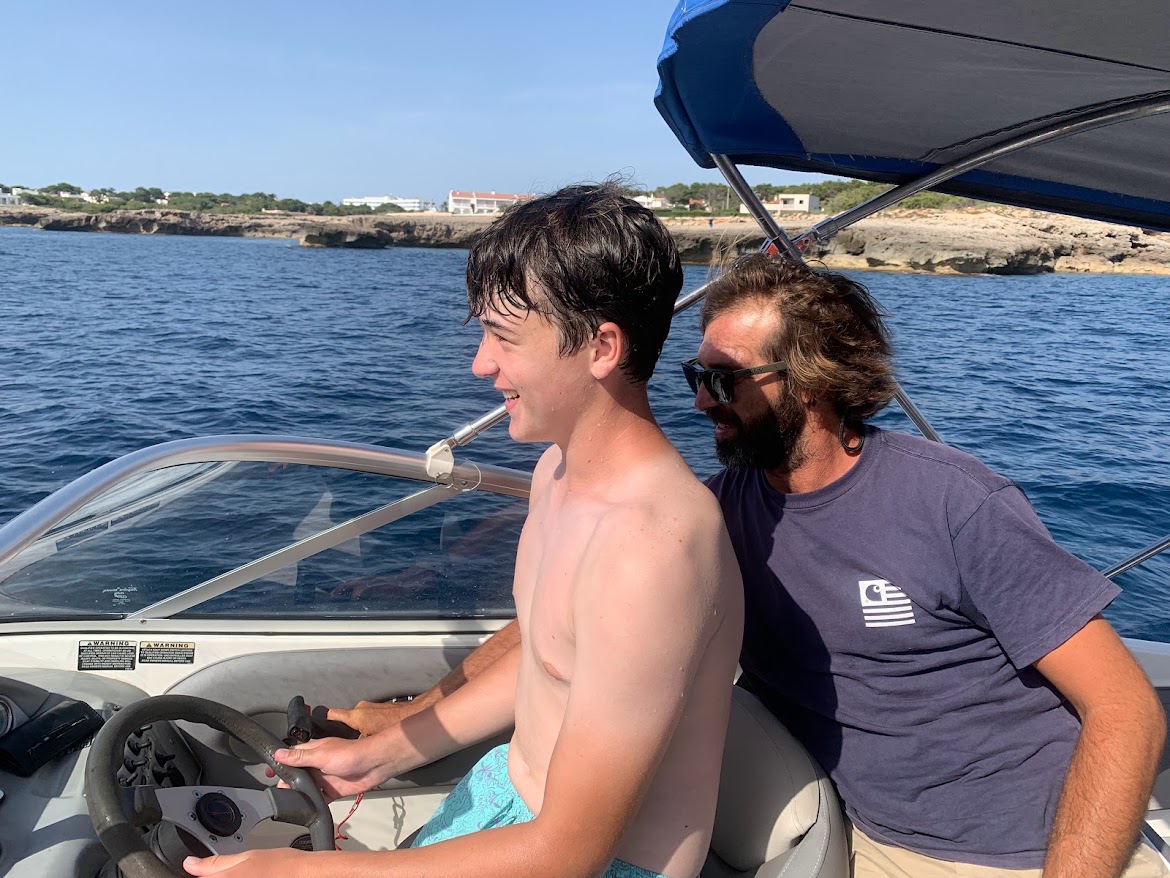 A teenager steering the boat with an adult beside him, coastline behind