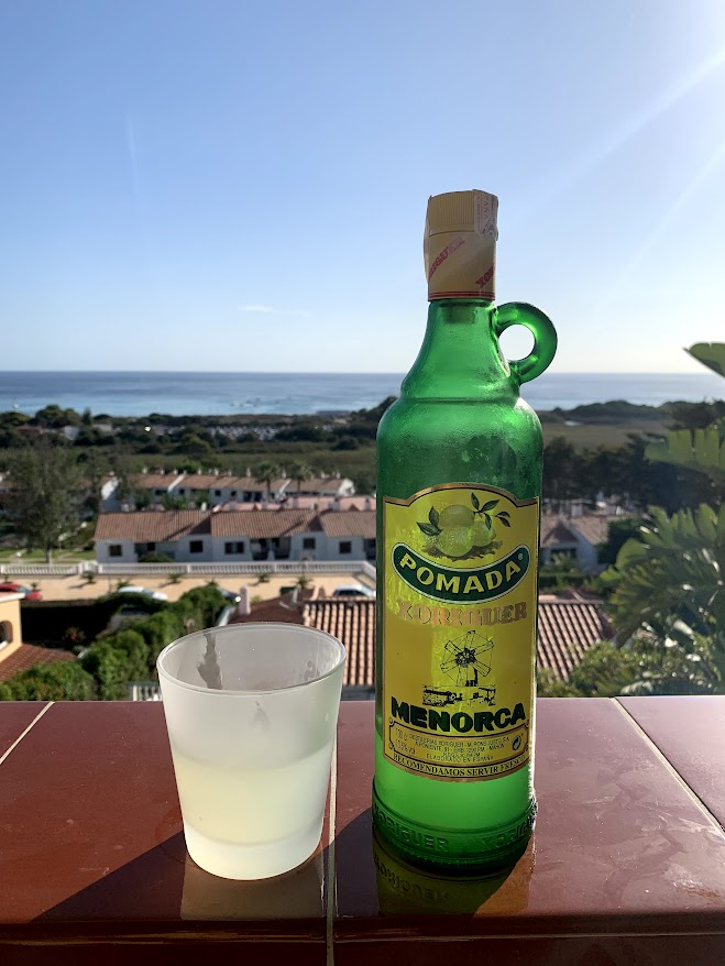 A bottle of Xoriguer Pomada and a glass, overlooking the coast